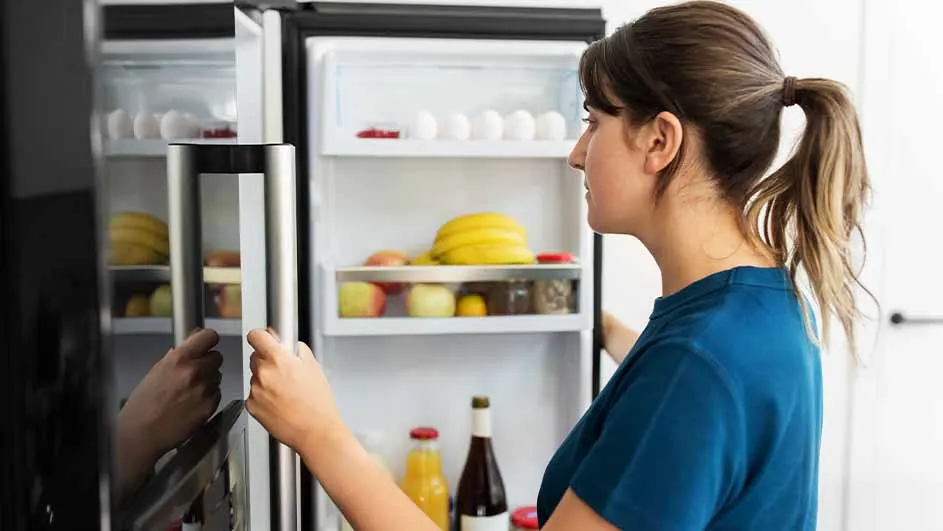 Vrouw in blauw shirt opent koelkast gevuld met eieren, fruit, en flessen.