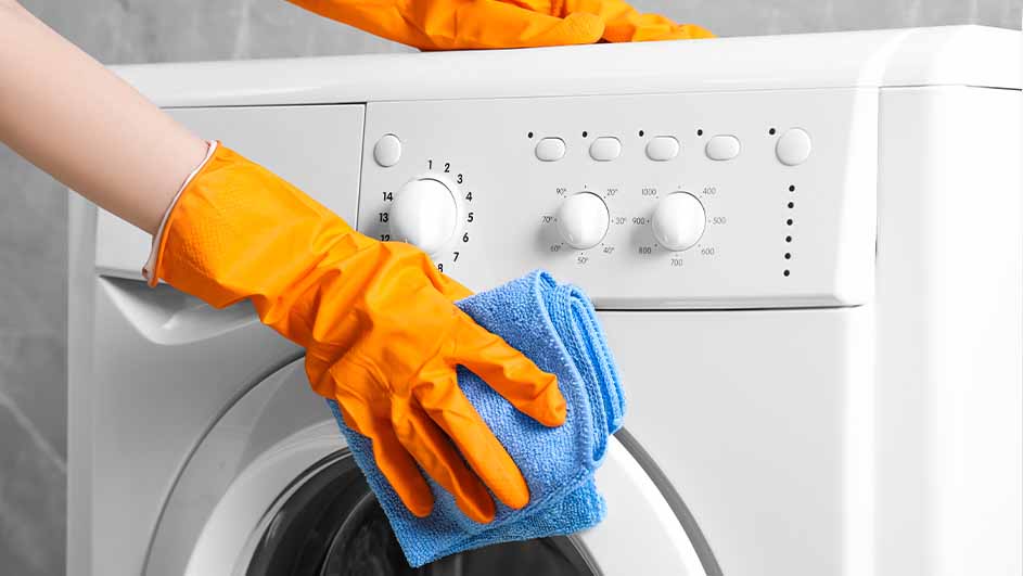 Een wit wasmachine wordt schoongemaakt met een blauwe doek en oranje handschoenen tegen een grijze achtergrond.