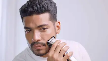 Een man gebruikt een tondeuse op zijn gezicht, tegen een witte achtergrond. Hij draagt een wit shirt.