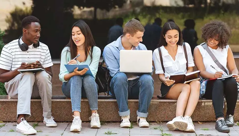 Vijf studenten zitten buiten op een trap en werken samen met notitieblokken, pennen en een laptop. Er zijn geen zichtbare merknamen.