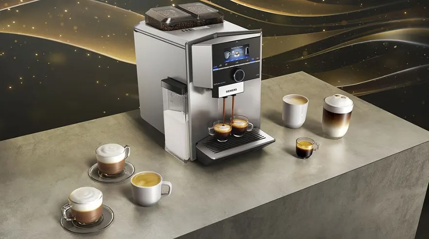 Een zilverkleurige Siemens koffiemachine zet koffie in twee glazen kopjes op een grijze tafel met zes andere kopjes.