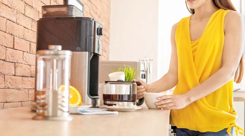 Vrouw in gele top schenkt koffie uit een koffiezetapparaat in een witte mok. Er staat een stenen muur en een glazen pot op het aanrecht.