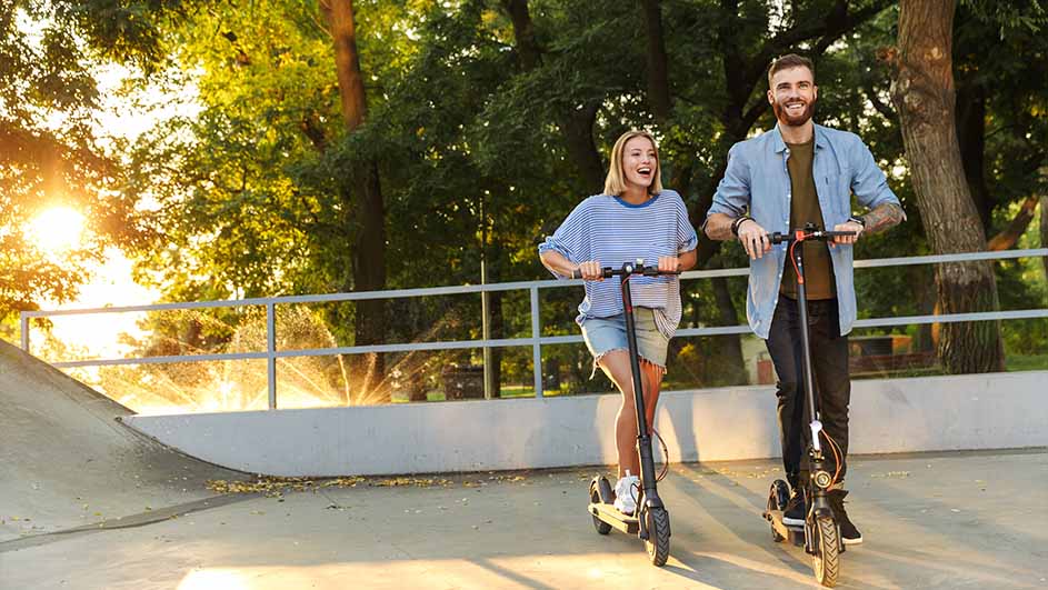 A man and a woman ride scooters in a park with green trees and a white fence.