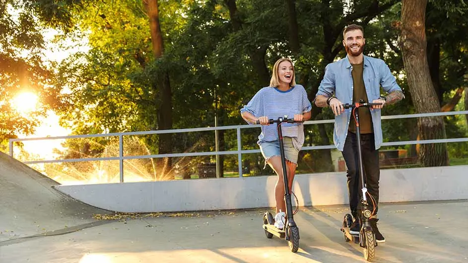 A man and a woman ride scooters in a park with green trees and a white fence.