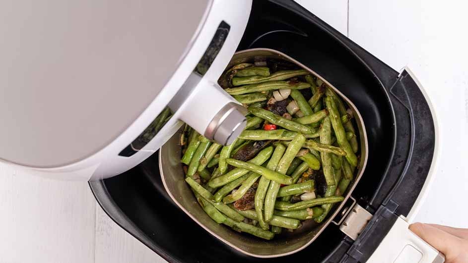 Green beans and other vegetables are in the air fryer basket. The white air fryer is on a white surface.