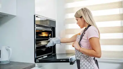 Een vrouw met een ovenhandschoen buigt zich voorover naar een open oven in een keuken.
