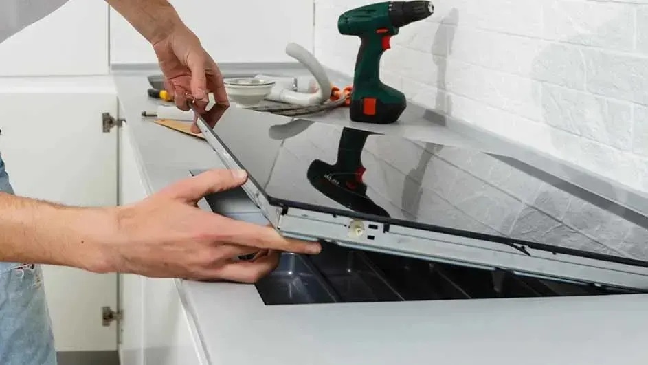 Monteur plaatst een glaskeramische kookplaat in het werkblad; gereedschap ligt klaar, waaronder een accuboormachine en kabels.