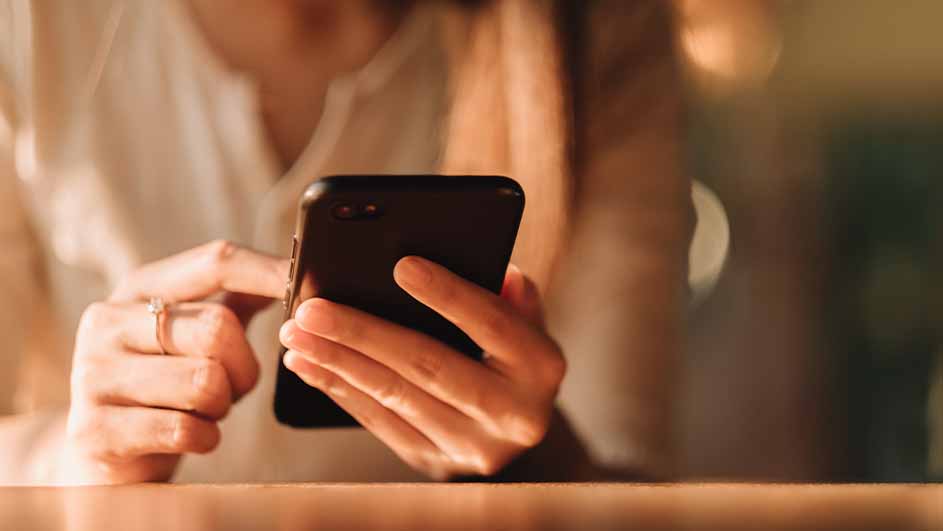 Een vrouw in een wit shirt gebruikt een zwarte smartphone aan een houten tafel.