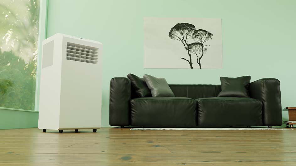 Witte airconditioner naast een zwarte bank met kussens, tegen een lichtgroene muur met een foto van bomen.