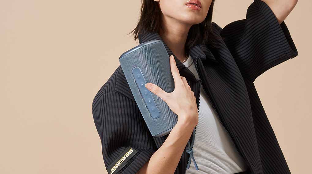 Een vrouw houdt een blauwe luidspreker vast, gekleed in een zwart jasje met witte top, tegen een beige achtergrond.