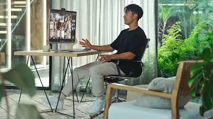 A man joins a video call at home, seated at a tidy desk with plants, soft daylight, and a calm, modern workspace.
