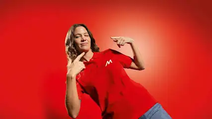 Vrouw wijst met beide handen naar zichzelf, draagt een rood poloshirt met een witte 'M' tegen een rode achtergrond.