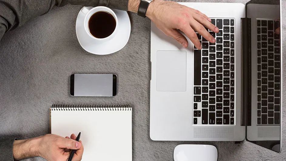 Een bureau met een laptop, kop koffie, notitieblok, pen, muis en telefoon op een grijze achtergrond.
