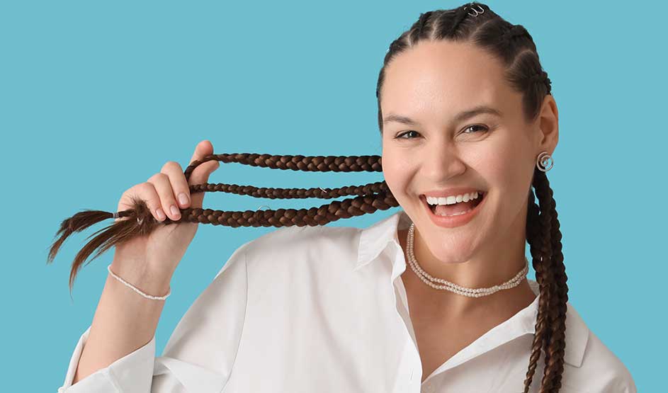 Een vrolijke vrouw met lange vlechten houdt haar haar speels vast en lacht breed tegen een egaal blauwe achtergrond.