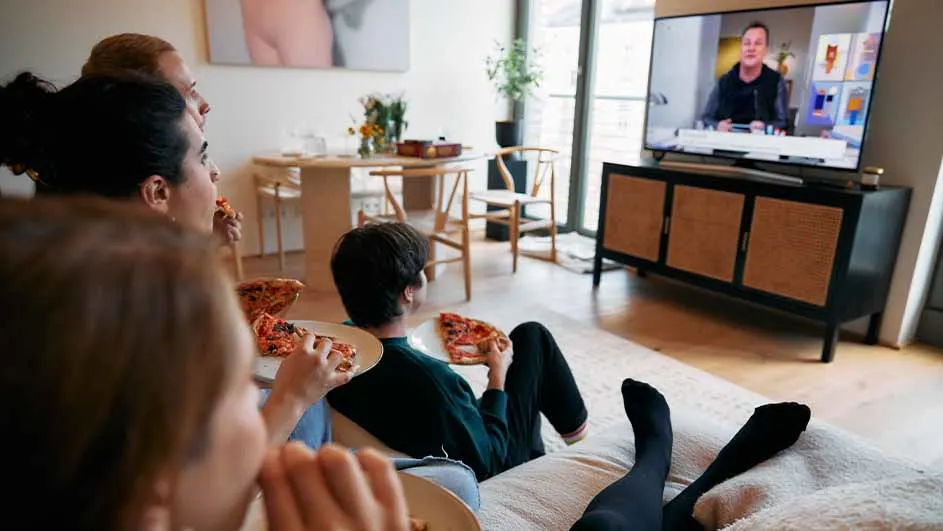 Een groep mensen eet pizza en kijkt tv in een woonkamer met een tafel en stoelen.