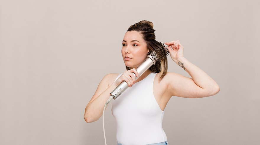 Een vrouw in een wit hemd gebruikt een zilveren föhnborstel om haar haar te stylen tegen een effen achtergrond.