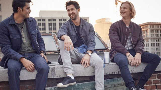 Drie mannen zitten op een stenen muur op een dak, lachend en in gesprek. Gebouwen zijn op de achtergrond te zien.