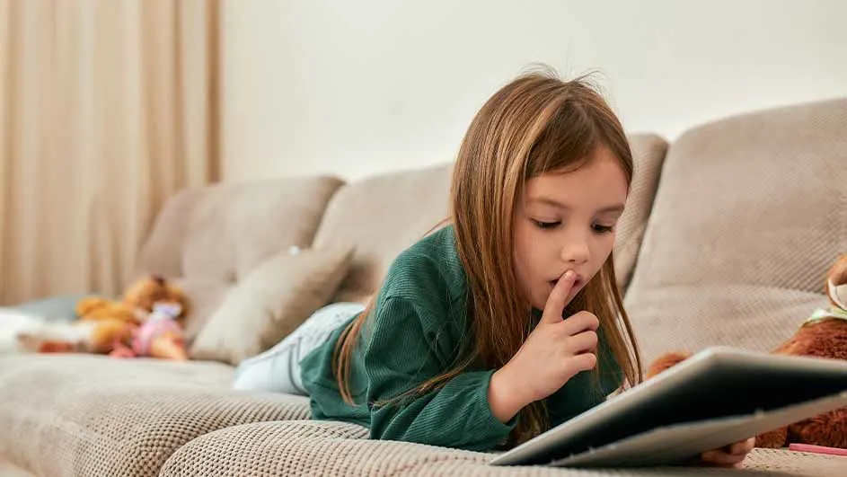 Meisje in groen shirt ligt op beige bank met tablet. Ze houdt een vinger voor haar mond. Knuffels op de achtergrond.