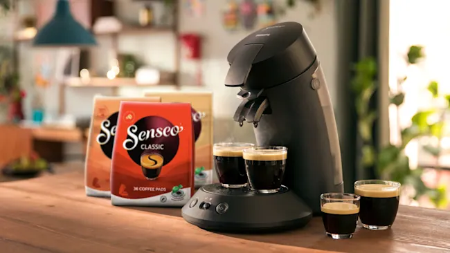 Een Senseo koffiezetapparaat met koffiepads en glazen koffie op een houten tafel, tegen een wazige achtergrond.