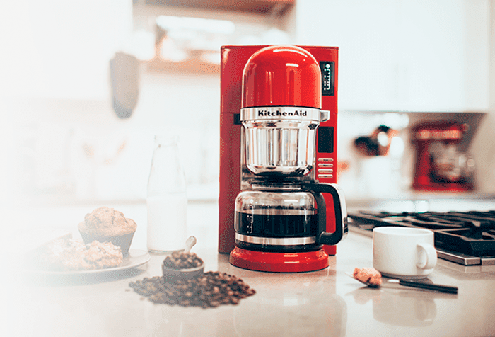 Een rode KitchenAid koffiemachine staat op een aanrecht, met een kopje, muffins, koffiebonen en een melkfles.