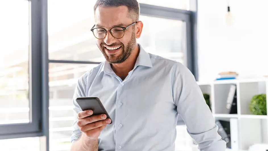 Een man met een blauw overhemd en een bril kijkt lachend naar een zwarte telefoon in een kantoor.