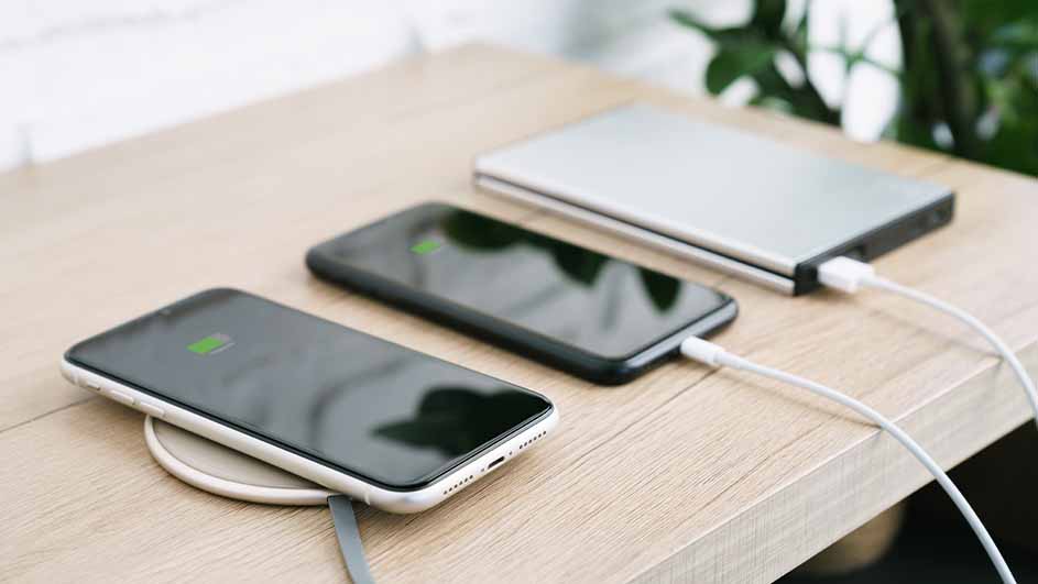 Twee telefoons en een powerbank liggen op een houten tafel. De telefoons tonen een groene batterij-indicator. Witte kabels verbinden apparaten.