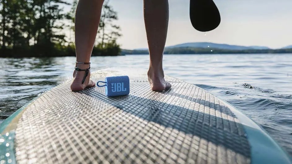 Een persoon staat op een supboard op het water, met een blauwe JBL speaker aan de zijkant. Een peddel is zichtbaar.