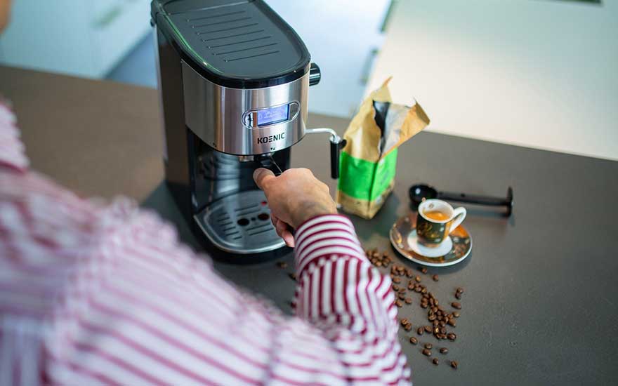 A man in a striped shirt uses a KOENIC espresso machine, with scattered beans and a tiny cup of espresso on the worktop.
