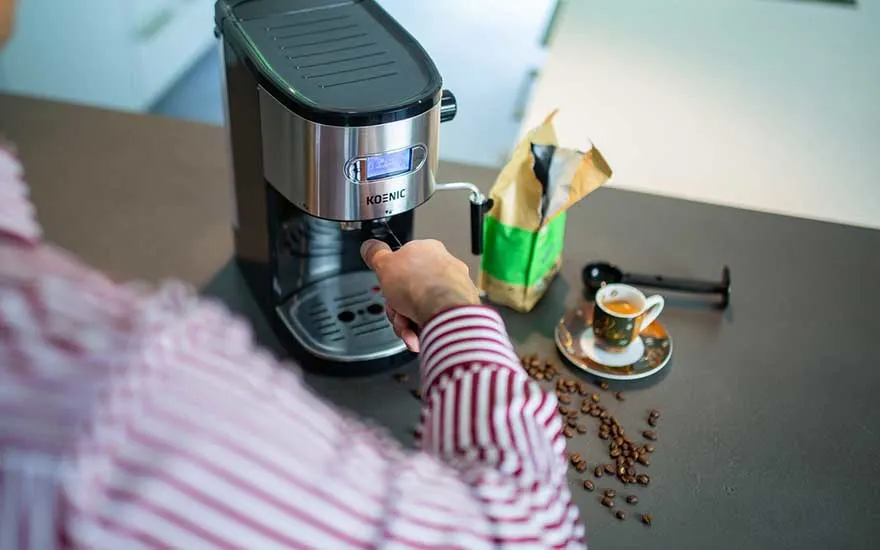 A man in a striped shirt uses a KOENIC espresso machine, with scattered beans and a tiny cup of espresso on the worktop.