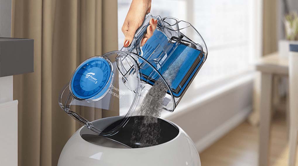 Een hand leegt een doorzichtige stofzuigercontainer met blauw filter in een witte afvalbak. Op de achtergrond gordijnen en een tafel.