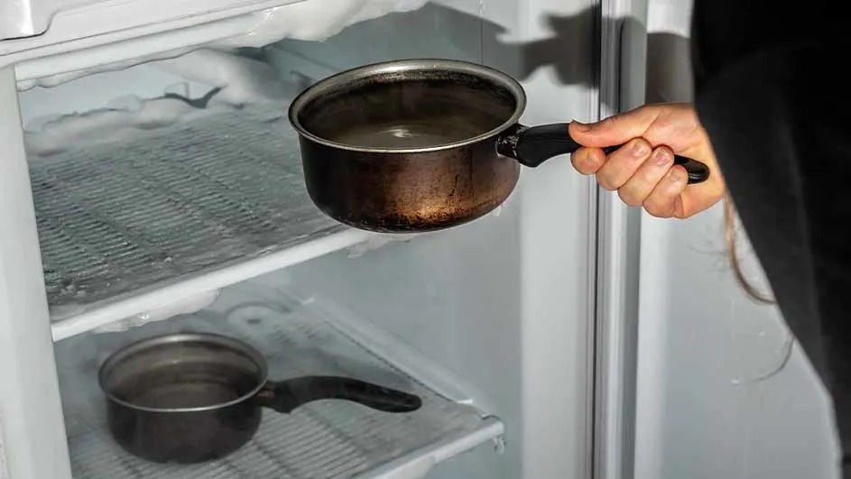 A hand holds a pot in front of an open freezer. The freezer has ice buildup and another pot on a shelf.