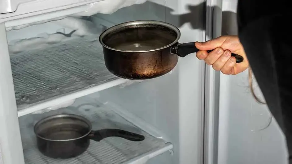 A hand holds a pot in front of an open freezer. The freezer has ice buildup and another pot on a shelf.