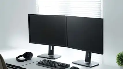 Twee zwarte monitoren staan op een wit bureau, met een toetsenbord, muis en hoofdtelefoon. Achtergrond: wit met witte jaloezieën en een plant.