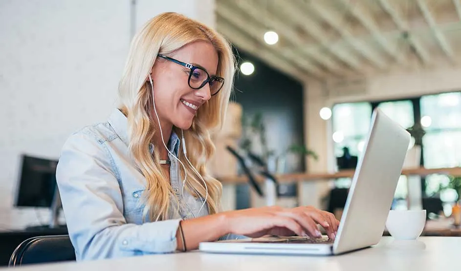 Blonde woman with glasses and wired earphones smiles at her laptop in a bright, modern office, coffee cup beside her.