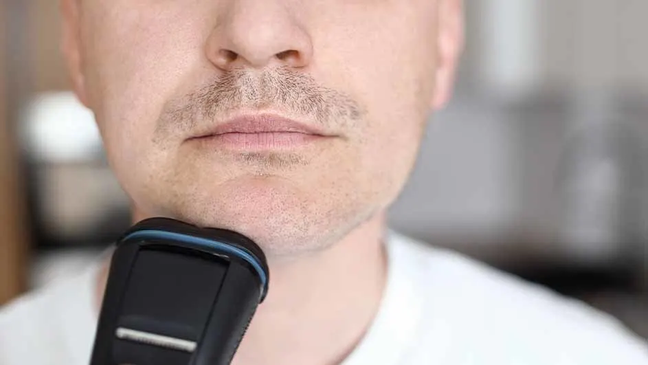Een man scheert zijn gezicht met een zwart elektrisch scheerapparaat. De achtergrond is vaag en licht.