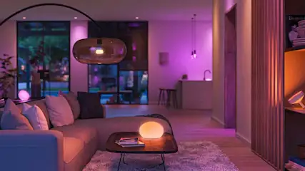 Woonkamer met bank, tafel, lampen en een open keuken op de achtergrond, verlicht in paarse en oranje tinten.