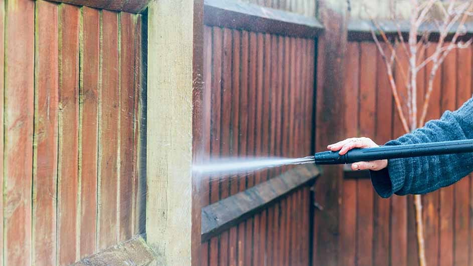 A person uses a Kärcher pressure washer to clean a wooden garden fence, making the wood look fresher and brighter with the water jet.