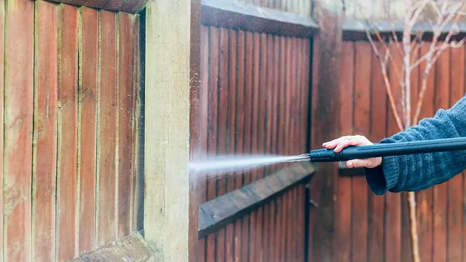 A person uses a Kärcher pressure washer to clean a wooden garden fence, making the wood look fresher and brighter with the water jet.