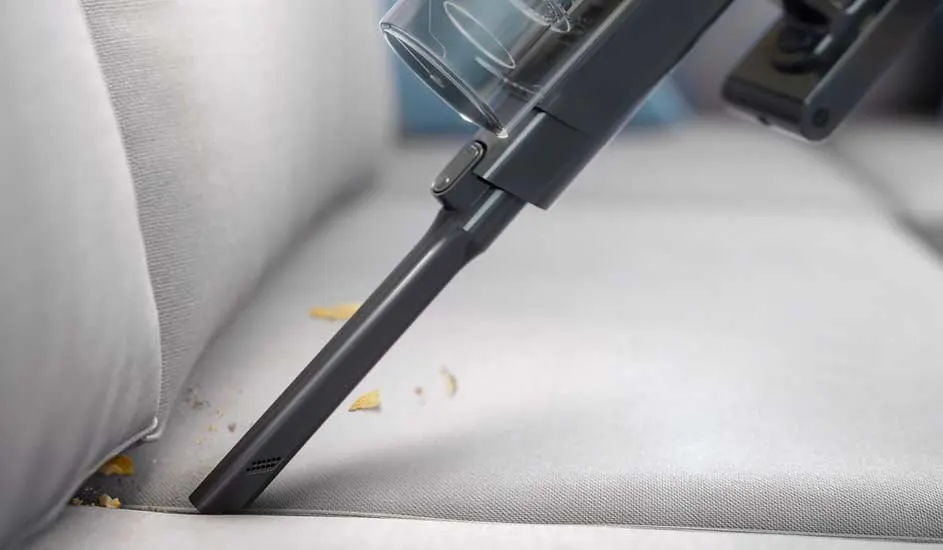 A slim vacuum nozzle cleans scattered crumbs from the pale fabric sofa seam, reaching deep between cushions with precision.