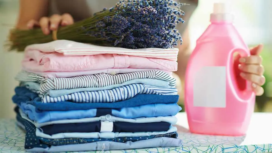 Gestapelde kleding met lavendel en een roze wasmiddelfles op een tafel.