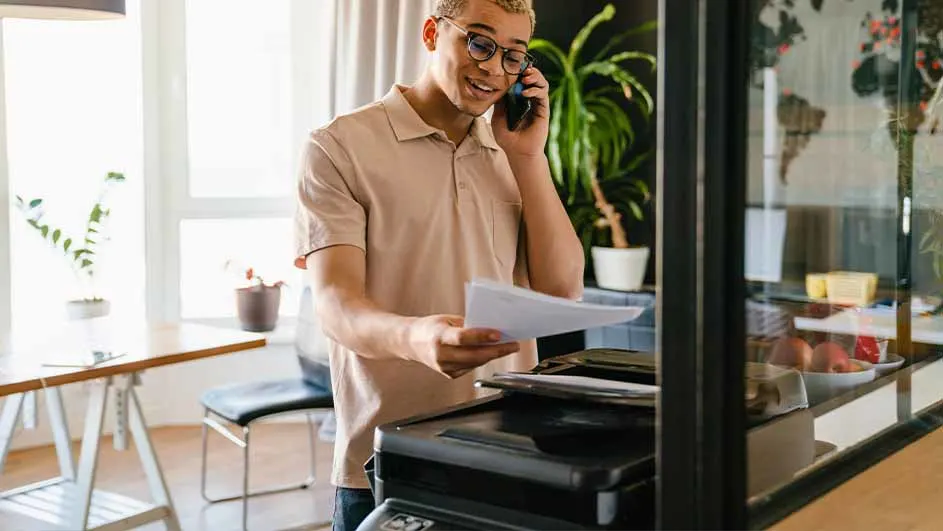 Man met bril aan de telefoon, houdt papier boven printer. Achtergrond: planten, tafel en stoel.