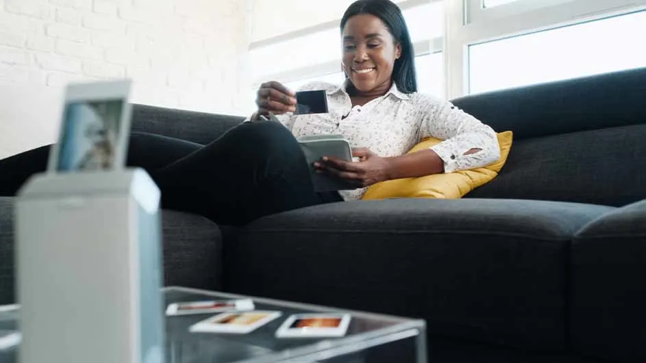 Vrouw zit op een zwarte bank met een gele kussen, houdt een tablet en creditcard vast. Op de tafel liggen foto's.
