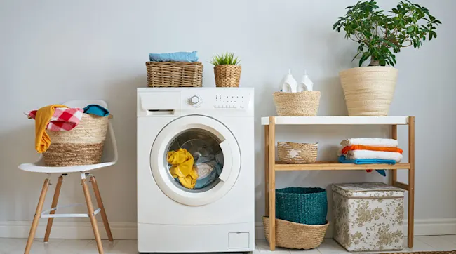Laundry room with a white washing machine, baskets with towels, a plant, and a wooden shelf.