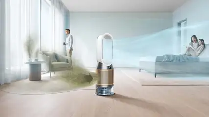 In een lichte kamer staat een luchtzuiveraar, die lucht blaast naar een bed met twee lezende personen. Een man staat bij een raam.