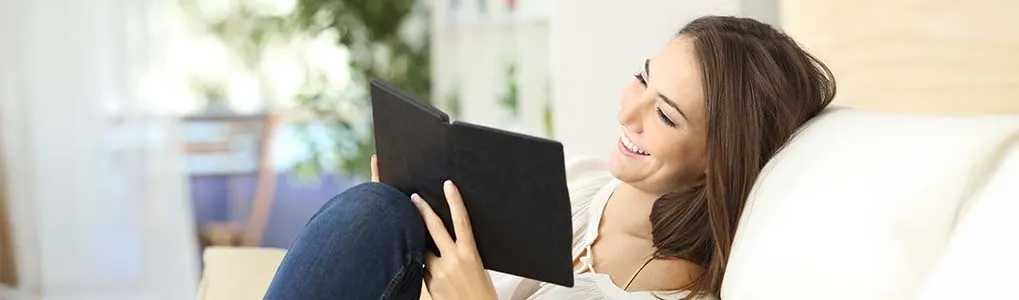 Een vrouw ligt ontspannen op een lichte bank en leest met een glimlach op haar gezicht op een zwarte e-reader. De achtergrond is licht en huiselijk, met zachte kleuren en een vage plant in beeld.