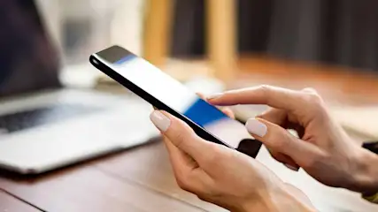 Vrouw gebruikt een zwarte smartphone met een vinger. Op de achtergrond een laptop op een houten tafel.