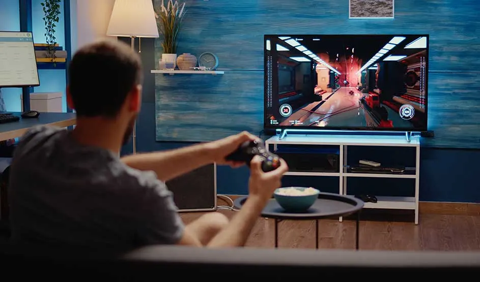Een man speelt een videogame met controller voor de tv in een gezellige woonkamer met blauwe muur.