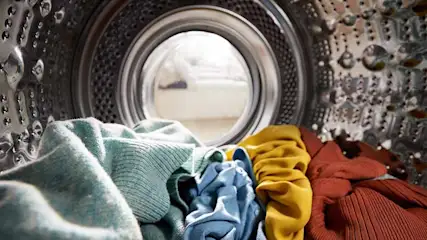 Inside a silver washing machine, colorful clothes are seen through the door. Garments in blue, yellow, and red are visible.