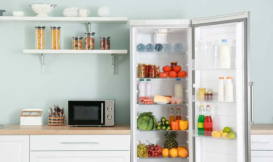 Een lichte keuken met open koelkast vol groenten, fruit, flessen en potten, plus planken, wit servies en een magnetron op het werkblad.