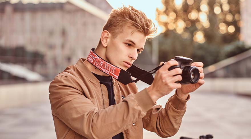 Een man in een bruine jas houdt een camera vast met een rood-witte band, op een vage achtergrond van gebouwen en bomen.